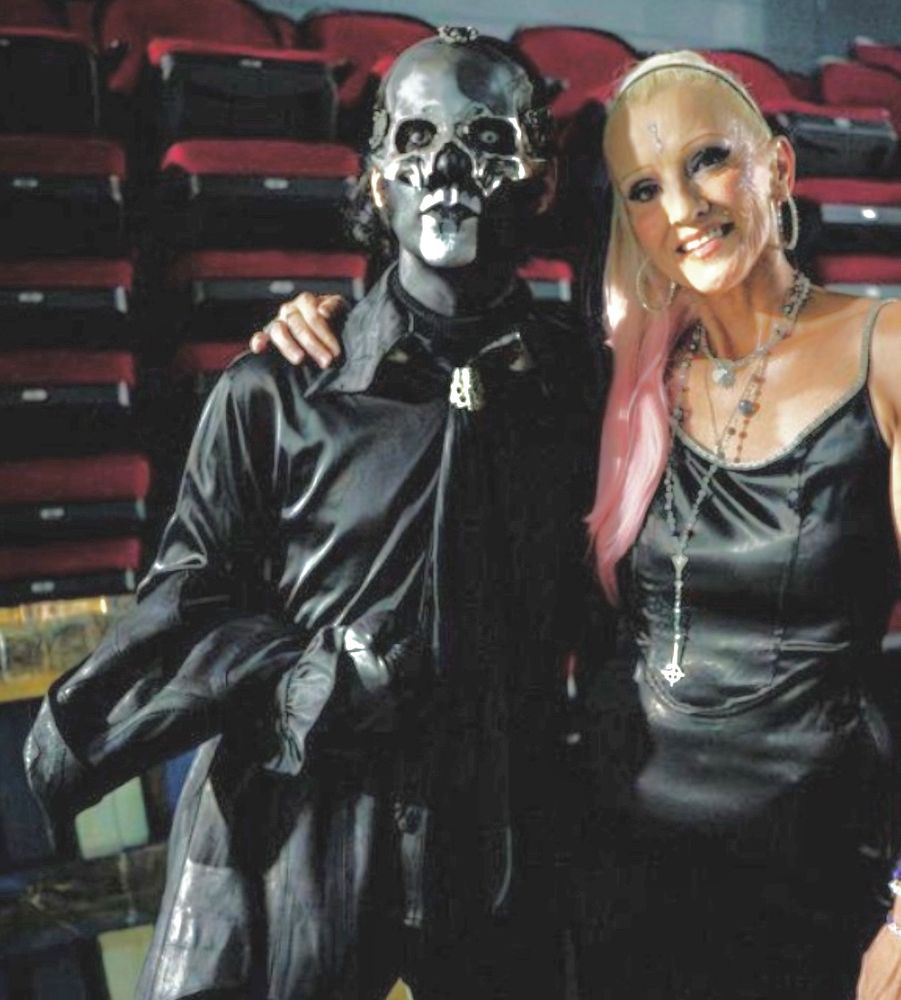 Papa V Perpetua and Vanessa Warwick posing for a photo off stage. Papa is carrying his leather jacket while staring straight at the camera, while Vanessa has her head tilted and smiling
