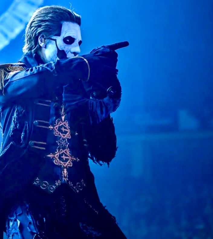 Papa Emeritus IV points at something while performing. He is donning the military jacket ensemble in blue background and lighting