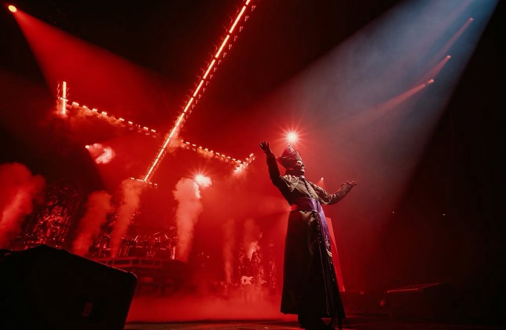 Papa V Perpetua in his Year Zero cassock outfit gesturing with both hands upwards to the unseen crowd. The lighting is red and ominous. 