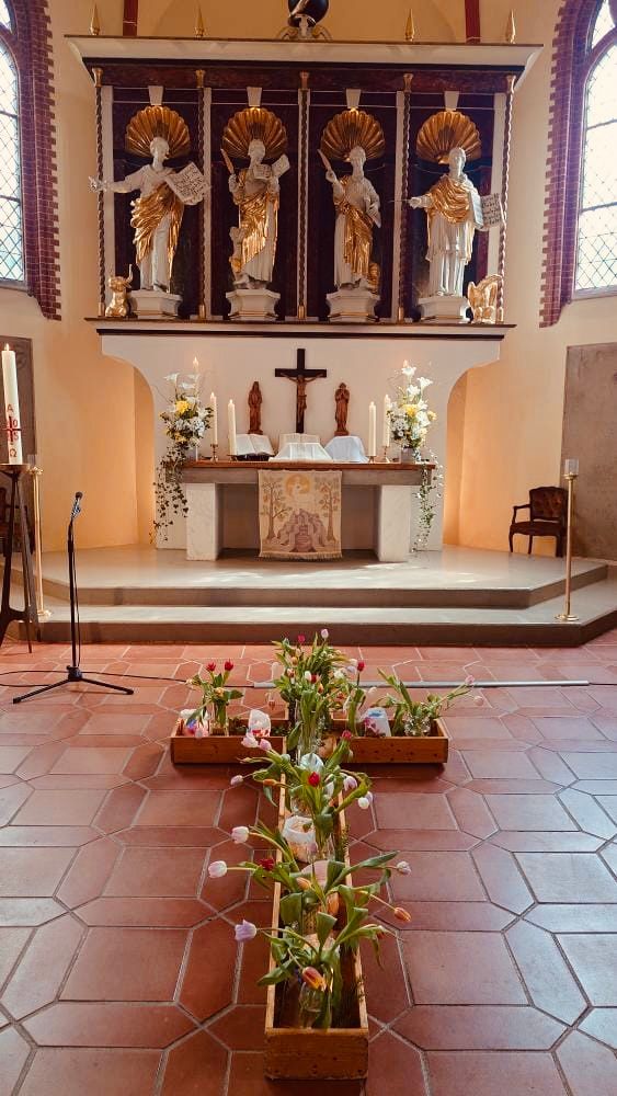 Bild einer Kirche, vor dem Altar ein mit vielen Blumen begrüntes gartengleiches Kreuz