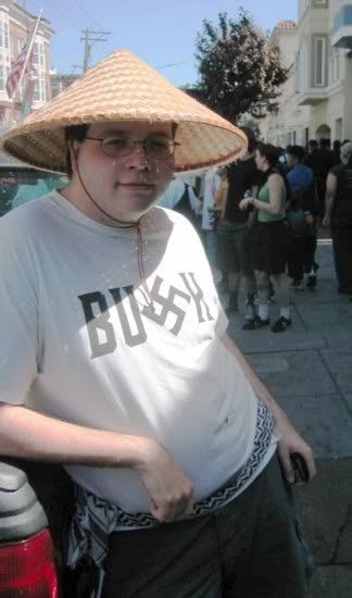 A young man in a conical hat wearing a white t-shirt with the word Bush written on it but the S is a swastika. I think he has a keffiyeh around his waist, but it looks like a knock-off pattern