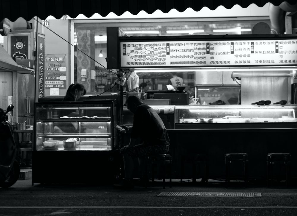 A man sits on a stool at a night market stall. His head is bowed as he uses his phone.
