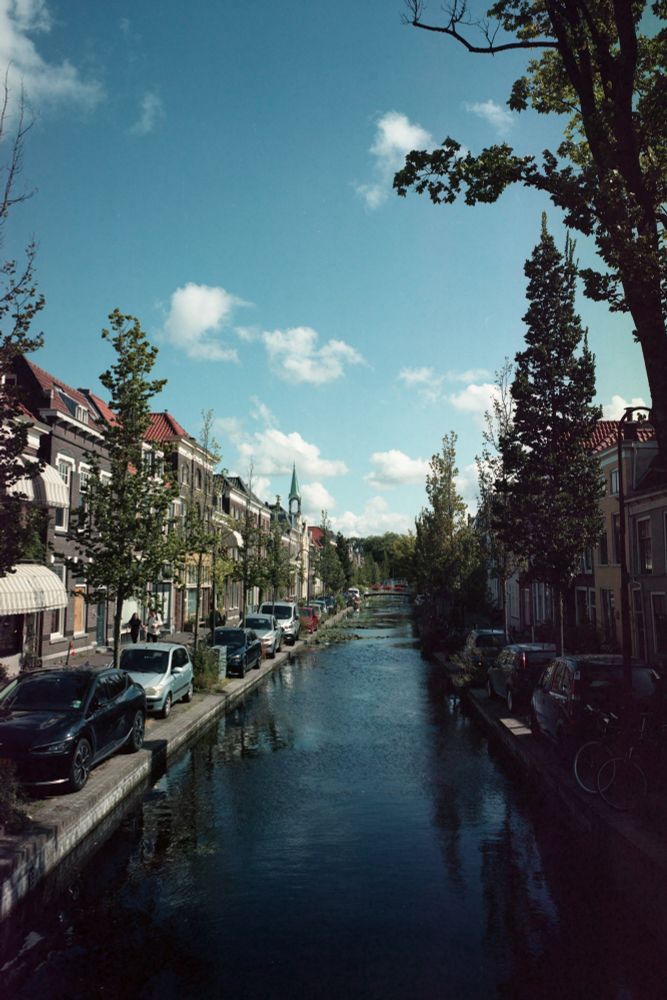 A painterly scene of a canal with cars parked next to it and old dutch houses behind trees. The sky is blue with a couple of clouds. Made with a Fuji GSW690ii on expired Kodak Portra 160.
