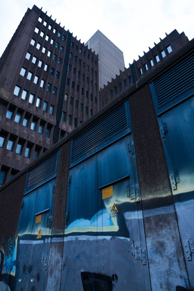 A dark concrete ominous looking building and an electricity substation in the foreground with blue graffiti