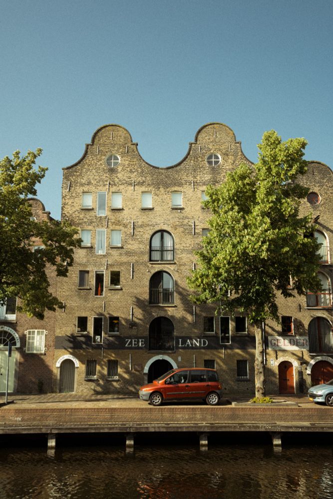 A Fiat Multipla parked on the edge of a canal, in front of an old building with the name Zee land painted on it