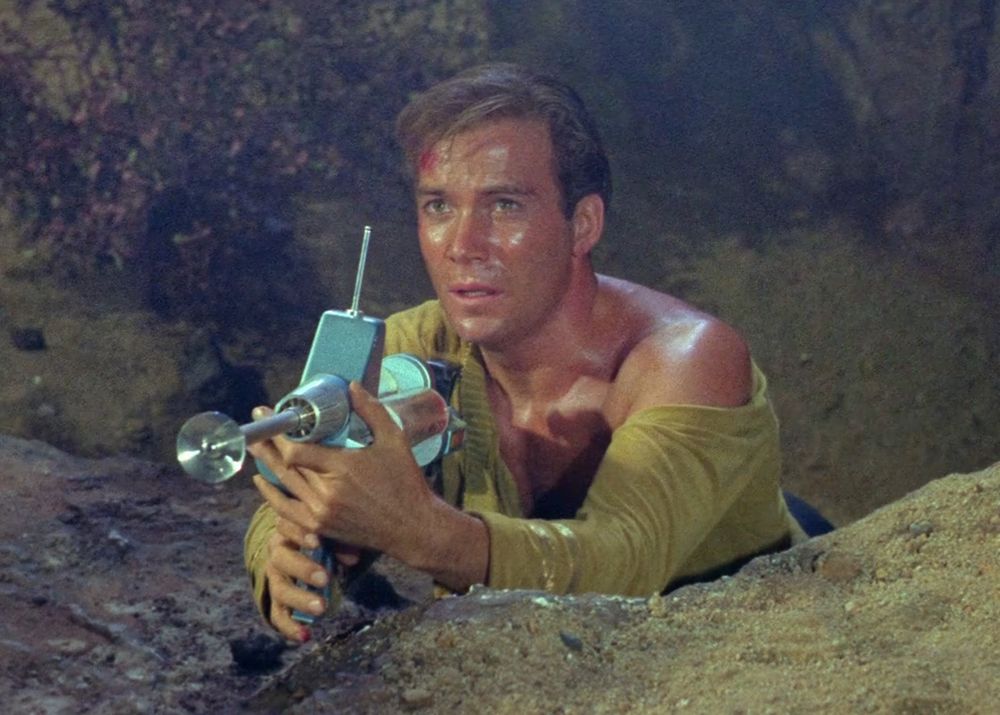 screencap of a scene from the pilot of "Star Trek: The Original Series." Captain James Kirk, played by a luminously beautiful young William Shatner, is crouching in like a giant pit of rocks, pointing THE most 1960s-looking ray gun rifle thing out of it. you see, Jim's college BFF goes mad with extrasensory power and Jim now has to try to kill him. it's a whole thing. that's not what is important. what's important here is that Kirk is sweaty, disheveled, with tousled hair and a few artful smudges of blood on his forehead, and he has inexplicably ripped his yellow uniform sweater in such a way that it looks from this shot as though he is wearing a cardigan with nothing under it and is letting it drop sexily off one shoulder. it's such a fucking look. they ONLY wore these uniforms in the pilot. after this they switch to a tunic with a stitched-on black collar that never budges, ever, so when Jim rips his shirt (which happens constantly) (well you follow me on my star trek alt you know that) the black collar never comes loose. it often gives him a kind of bondage look I'm not necessarily mad at, but the whole shirt does not absolutely come open like this, and that's a shame because he has extremely beautiful shoulders/neck/collarbone/chest action going on