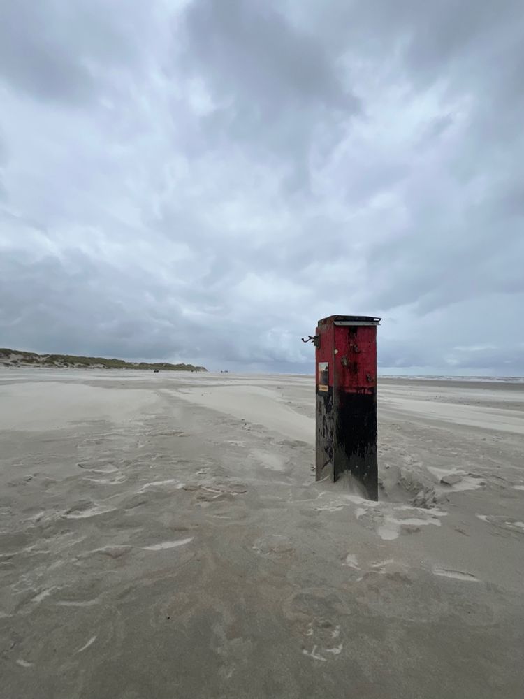 Een verlaten strand onder een bewolkte hemel, met een verweerde rood-zwarte houten paal op de voorgrond. De paal staat deels in het zand en draagt een klein, onleesbaar bordje. Links liggen duinen, rechts de zee. De scène straalt stilte en rust uit.
