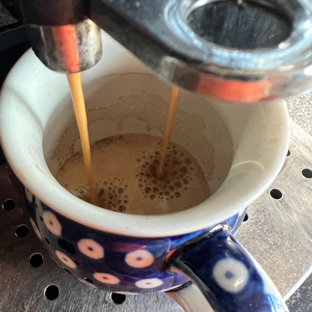 Koffie wordt gebrouwen, twee straaltjes koffie in blauwe mok met crema, op metalen lekrooster.