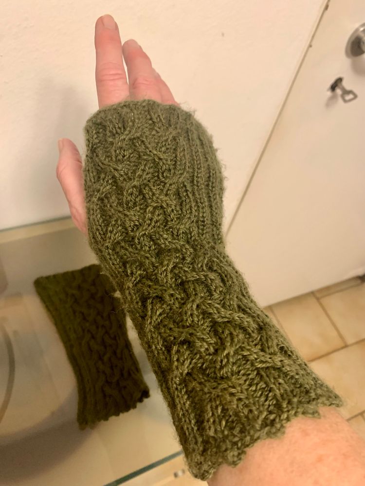 Eine Habd in einem olivgrünen Hangelenkswärmer mit Zopfmuster, gestrickt von der wunderbaren @reloaded2008 