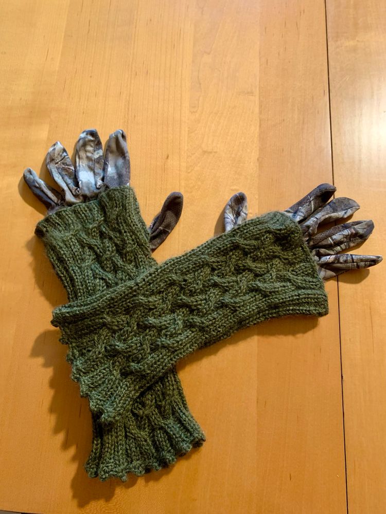 Ein paar gestrickte Handgelenkswärmer in olivgrün, darunter ein paar Fingerhandschuhe mit Tarnmuster