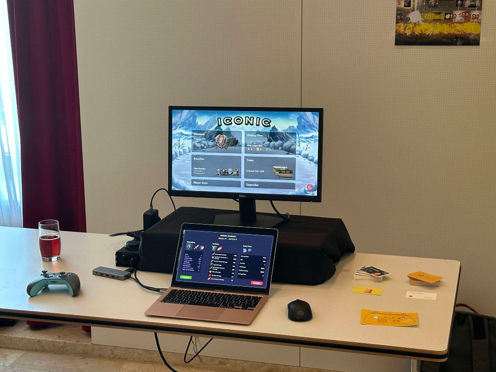 A table with a laptop, an external monitor, a game controller, a mouse, and a glass of red beverage. The monitor displays a game menu titled 'ICONIC'. The laptop shows the game's user interface. Business cards and a yellow flyer with a gamepad icon are also on the table.