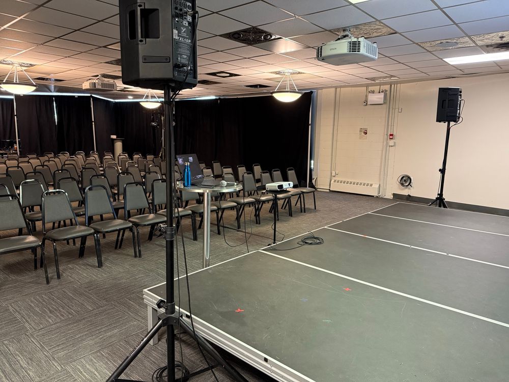 A photo taken offstage from stage left, showing the seats where up to 85 audience members can sit to watch Calgary Fringe Festival shows, of which mine is one. Two speakers flank the stage, and a table holding a laptop, a microphone, and a water bottle are visible. There’s is a projector centre stage.