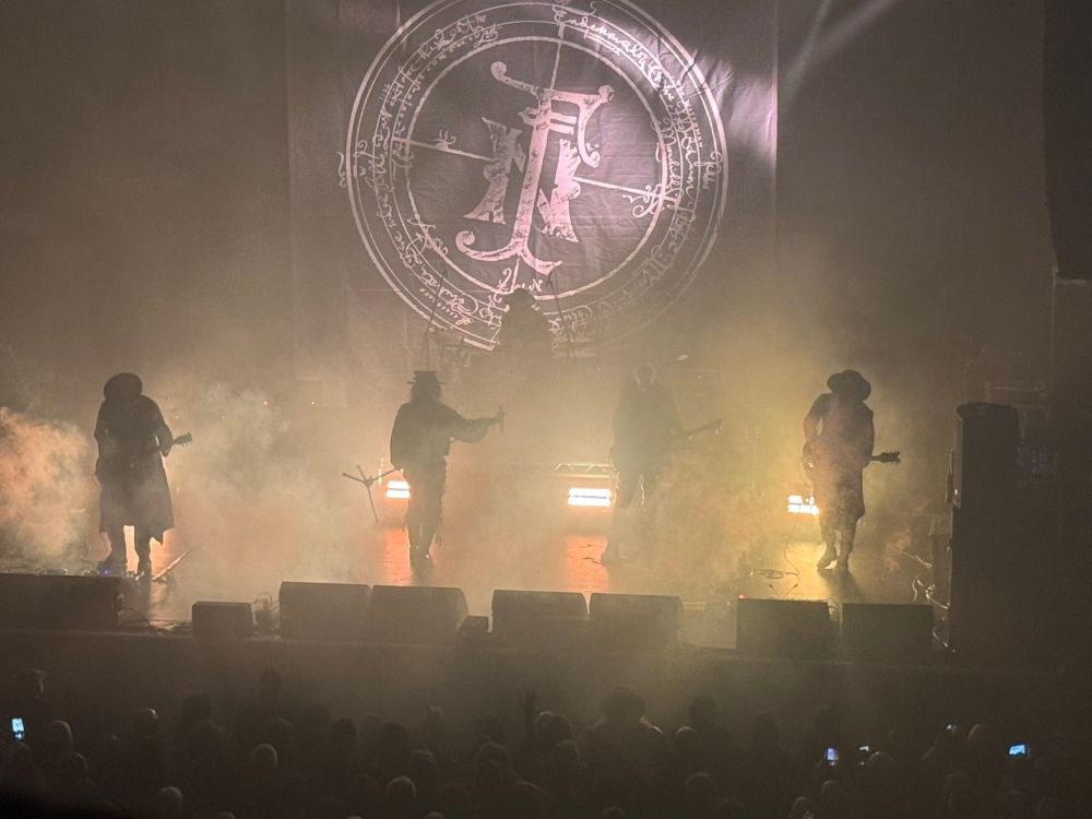 Four silhouetted figures on a stage full of smoke with a gothic logo above, the band Fields of the Nephilim performing live 