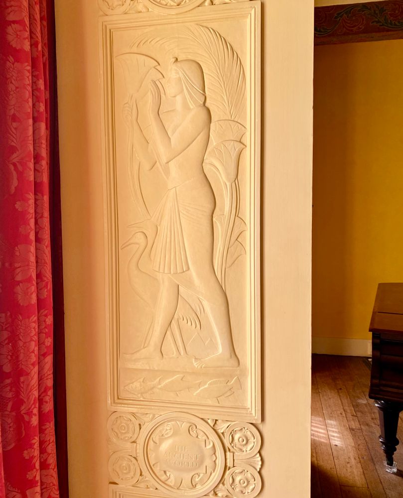 A wall moulding showing a male Egyptian-esque figure, captioned as ‘The Ancient World’. The proportions of the figure’s body may suggest it was inspired by art of the Amarna period.