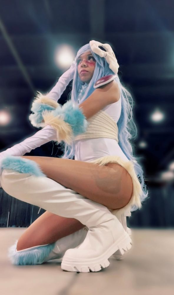 A cosplayer crouched and dressed as Nelliel wearing a white bodysuit and knee high boots. Both the body suit and boots are lined with teal and cream colored fur. She has matching gloves. Their hair is light blue and they’re also wearing red shoulder armor. A small skull mask rests on the side of their hair. 