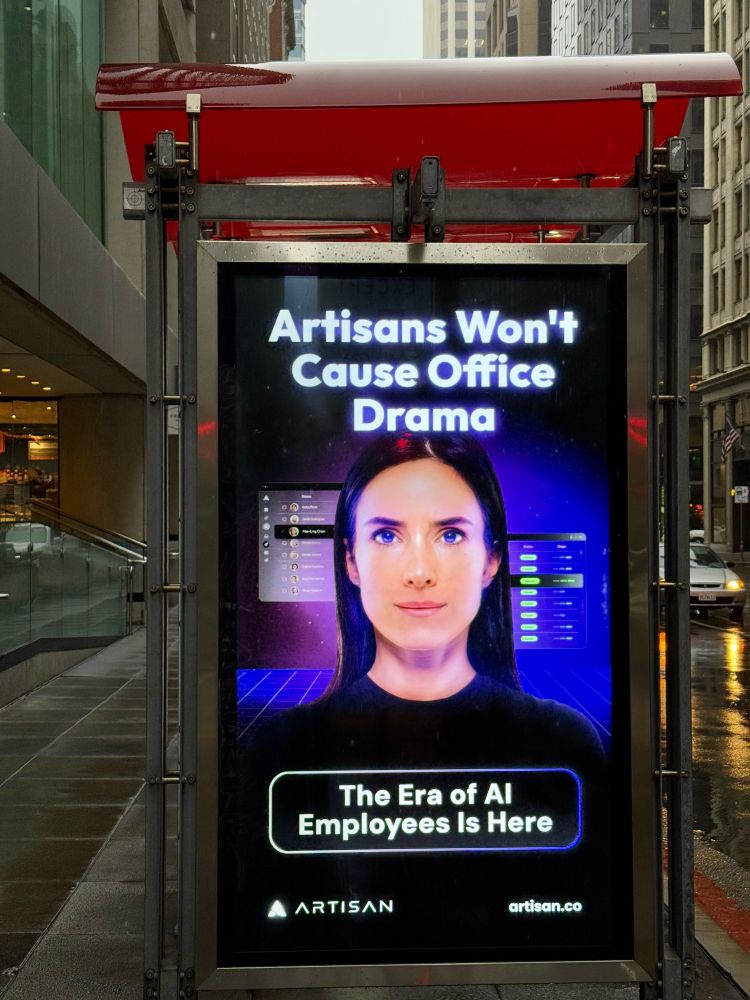 Advertisement at a bus stop: a creepy digital image of a woman’s face against an illuminated purple futuristic background. Text above the face reads: “Artisans Won’t Cause Office Drama” and below: “The Era of AI Employees Is Here” / ARTISAN / artisans.co