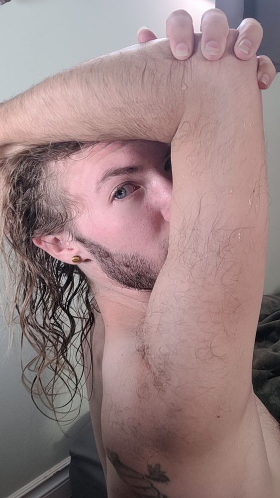 I'm stretching my shoulder above my head and looking back at the camera. My hair is damp from a shower and beads of water can be seen on my upper arm. 