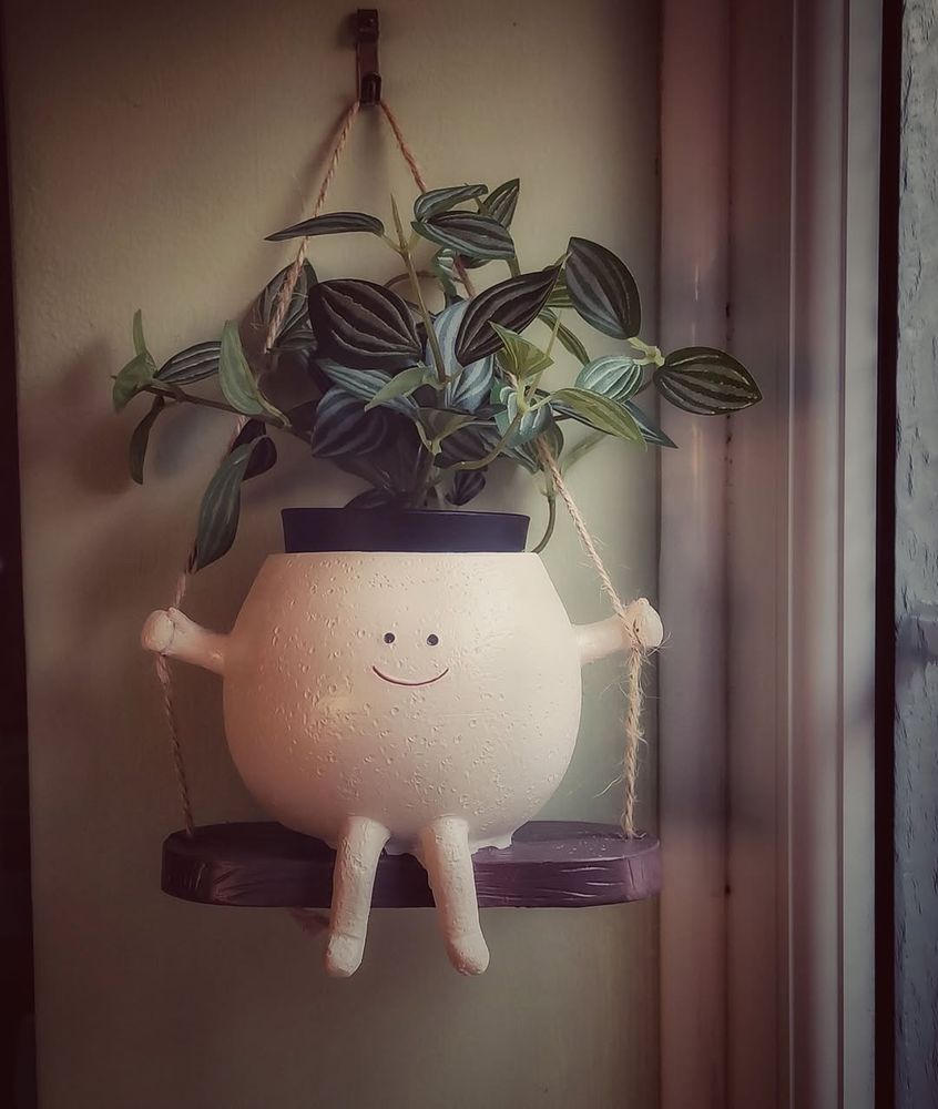A plant pot with smiling face, sat on a swing, containing a plant that makes it look like some crazy hairstyle.
