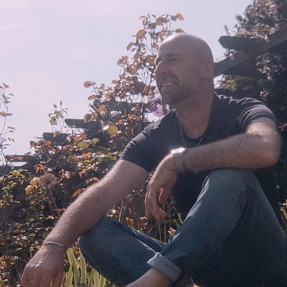 Man sat in garden in T shirt, in the UK enjoying warmer weather of the summer months.
