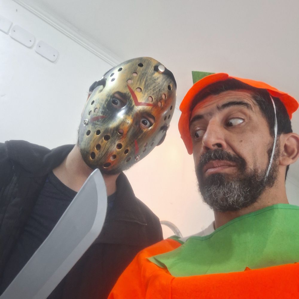 Eu vestido de abóbora ao lado do Delta, fantasiado de Jason. Ele segura um facão, para o qual eu olho com cara assustada.