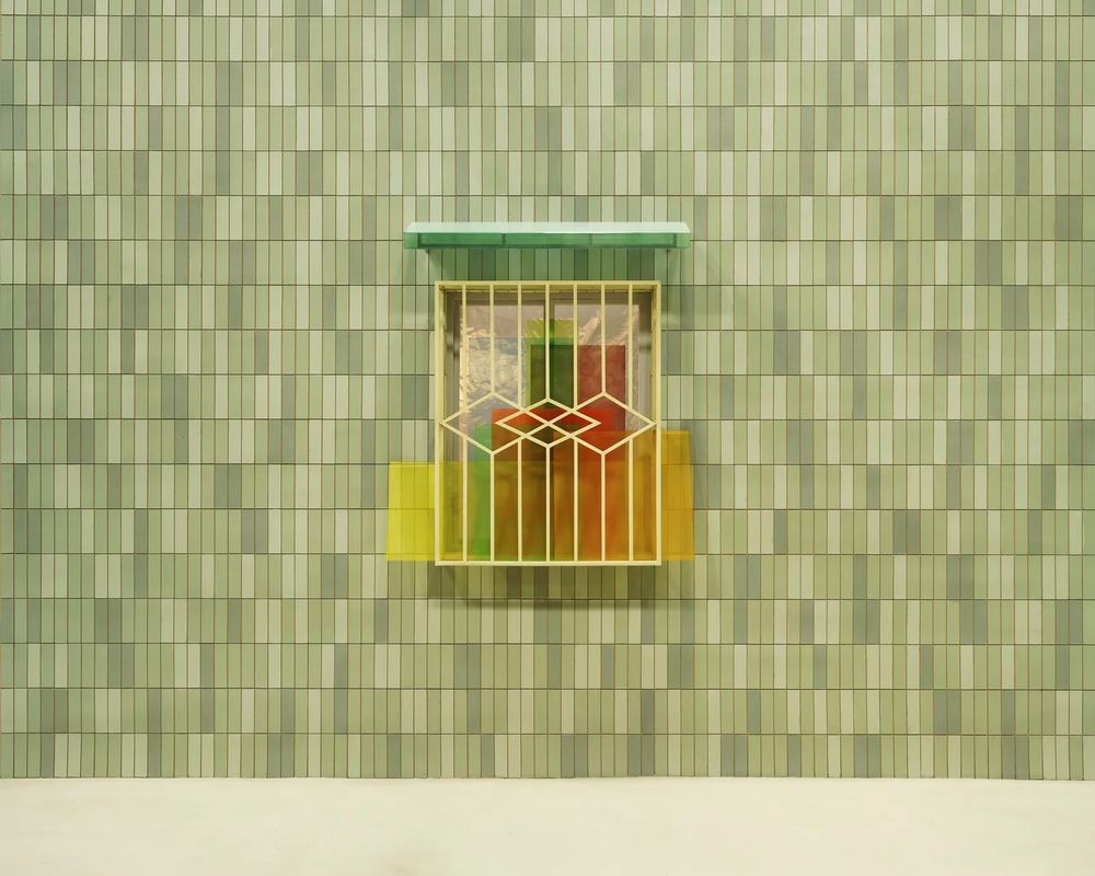 A small window with a slim green canopy, in a varied green tiled wall is covered with clear plastic sheeting serving as replacement glass. In front of a simple grill, rigid plastic pieces in yellow, red & green overlap to make a Mondrian type pattern.
