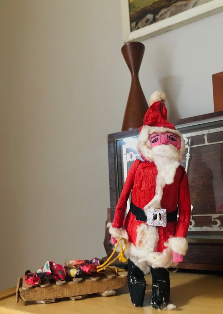 A homemade worn & battered Father Christmas made from a toilet roll, crepe paper & cotton wool dragging a lolly stick sleigh with tiny presents.
He looks like his life is a misery. 