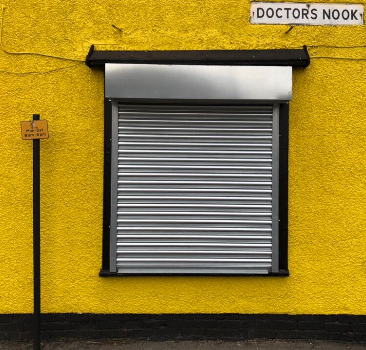 An acid yellow pebbledash wall with black painted bottom strip. A large square shuttered window with black frame centrally & a black vertical parking sign pole. 
A street sign ‘Doctors Nook’ at the right hand side of the wall.