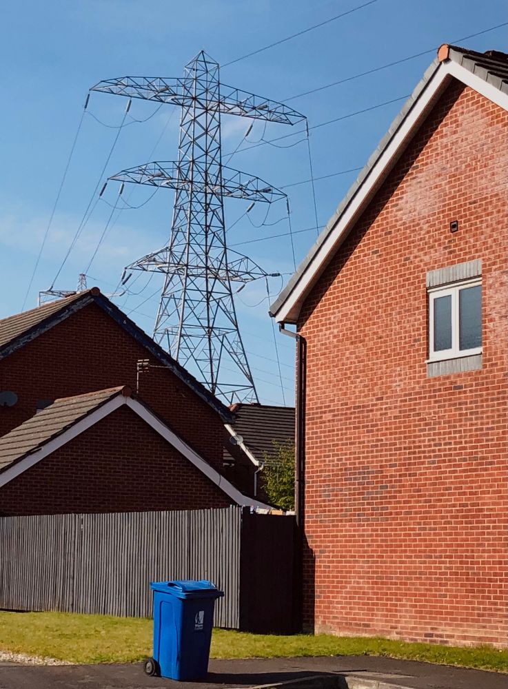 A solo bin on the pavement by the gable end of a red brick new build house. A long fence behind with another house. A large electricity pylon looms overhead.