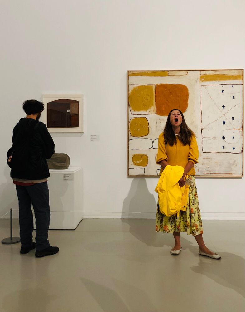 Inside a gallery a man in dark colours is looking at a small sculpture & wall piece. A woman in an ochre jumper holding a yellow raincoat has her back to a large William Scott abstract painting & is open mouthed in the middle of a big yawn.