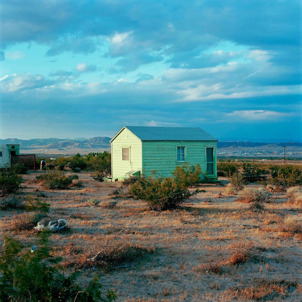 A small green wooden house sitting in the scrub of the desert. The mountains in the distance & a big pale blue sky. Low sunlight illuminates the side of the house.