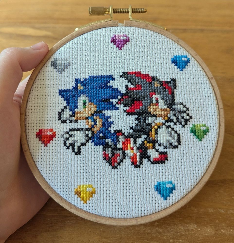 A cross stitch of the Sonic and Shadow pose from SA2 in pixel art form with 7 chaos emeralds surrounding them, framed in a 5 inch hoop frame