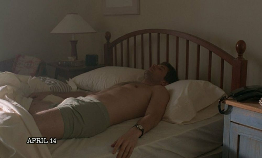 A still from The X-Files Season 2, Episode 25 "Anasazi." Agent Mulder is lying on his back asleep alone in a queen sized bed. He is a white man of average build with short brown hair and a 5 o'clock shadow. A white duvet cover is pushed down to his thighs. He wears a pair of light grey boxer briefs and an analog wrist watch on his left wrist. It's early morning so the room is dimly lit. There is a ray of light highlighting Mulder's bulge.