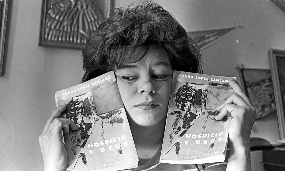 foto em preto e branco da escritora brasileira maura lopes cançado segurando seu livro "hospício é deus". ela tem um exemplar em cada mão e os pressiona contra o rosto. desvia o olhar, delineado, para a esquerda. tem cabelos curtos e ao fundo se podem ver algumas decorações.