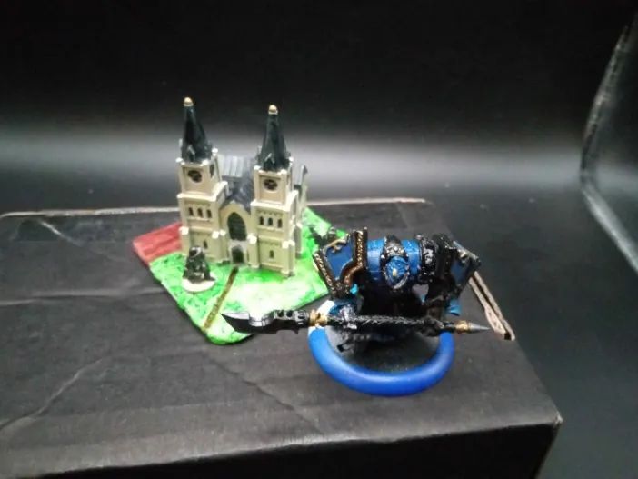 A model knight in front of a smaller-scale model church. The knight is in heavy blue armor with black and gold trim, carried a halberd in two hands, and looks rather like a Templar. The church is tan with a a black roof and green base, and some WW2 soldiers painted as "bronze statues" around its "landscaping".