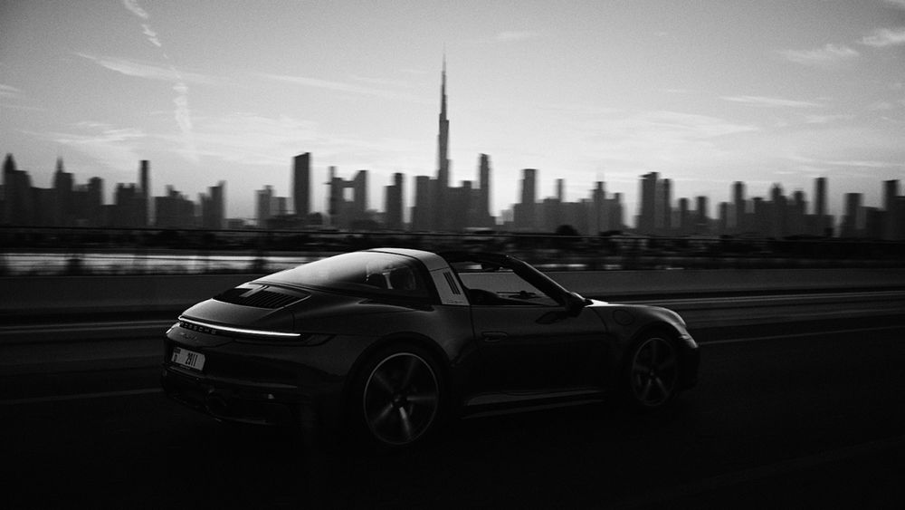 A black-and-white photograph of a Porsche 911 Targa 4S speeding along a road at dawn in Dubai. The car appears in motion with a smooth blur, capturing its elegance and speed. In the background, the iconic Dubai skyline stands against a soft, early morning sky. The image features a subtle grain, adding an artistic touch.