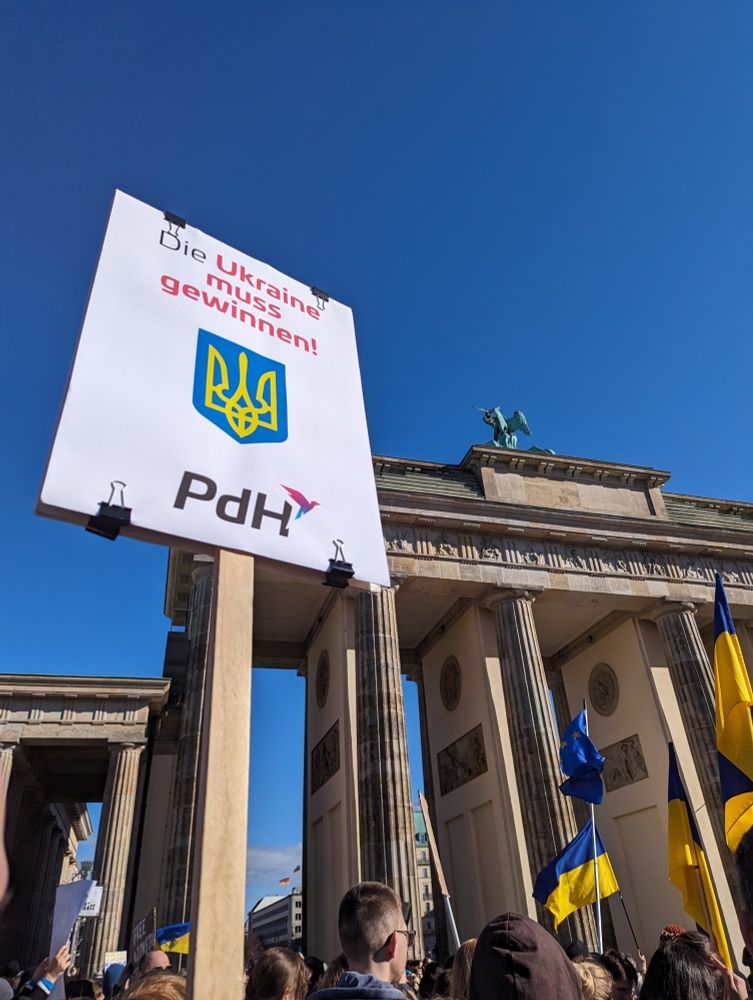 Foto von der Ukraine-Demo am Brandenburger Tor. Das Tor ist zu sehen sowie Ukraine-Flaggen und ein Plakat der PdH mit der Aufschrift "Die Ukraine muss gewinnen!"