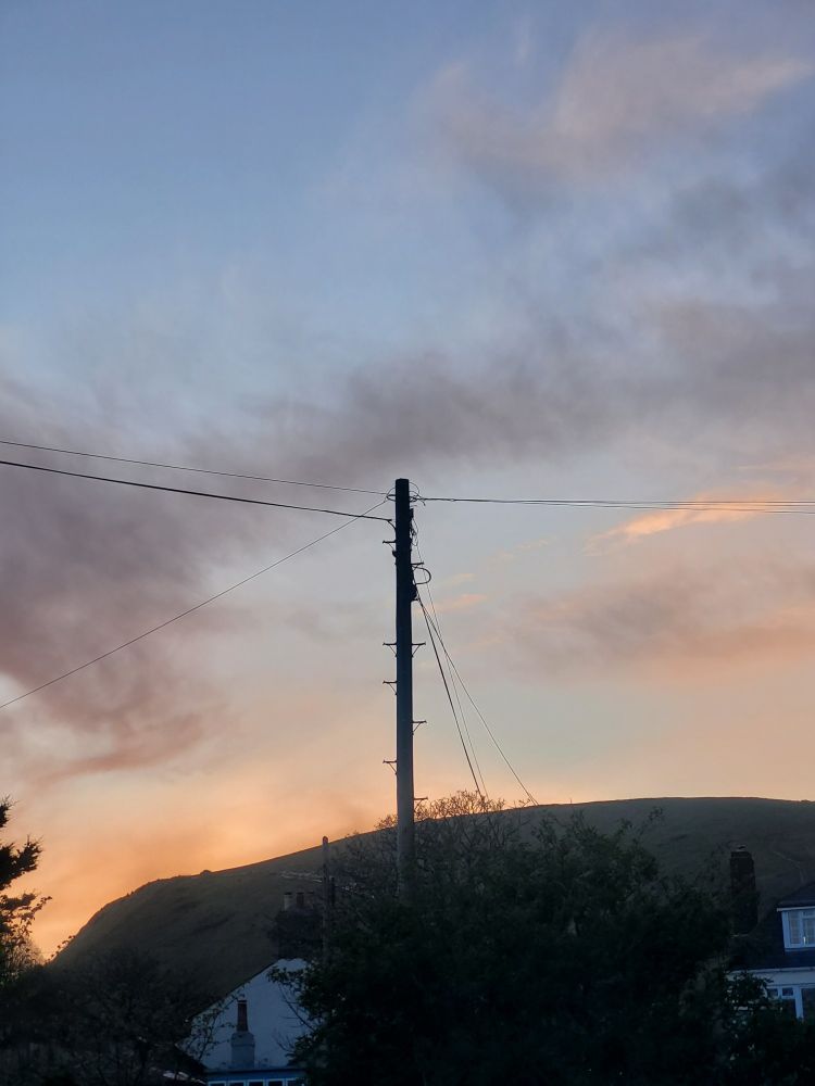 Telephone poll against sunset struck sky. A hill, 2 trees and 2 houses are in the lower background 