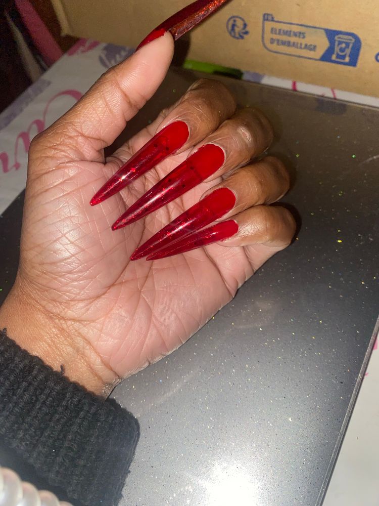 A picture of my left hand with four fingers folded on the palm with XXL stiletto Gel X tips with jelly red polish and red sparkles underneath 