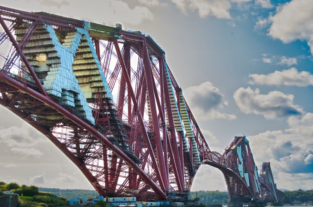 Die Aufnahme zeigt die Forth Bridge in Schottland, eine monumentale rote Stahlbrücke über den Meeresarm Firth of Forth. Ihre gewaltigen Fachwerkträger spannen sich in drei großen Bögen über das Wasser. Teile der Konstruktion sind eingerüstet und mit silbrigen Planen abgedeckt – sie wird gerade renoviert. Im Hintergrund leuchten blauer Himmel und weiße Wolken, unter der Brücke glitzert das Wasser, und am Ufer sind grüne Hügel zu erkennen.