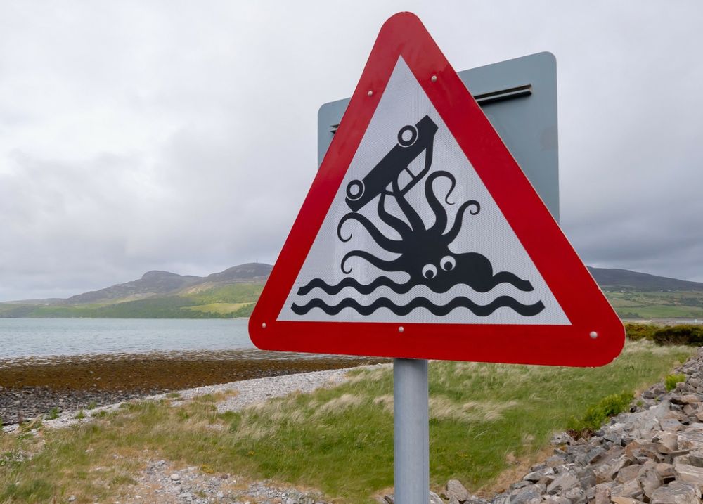 A triangular road sign depicting a car in the grasp of a large octopus with googly eyes.