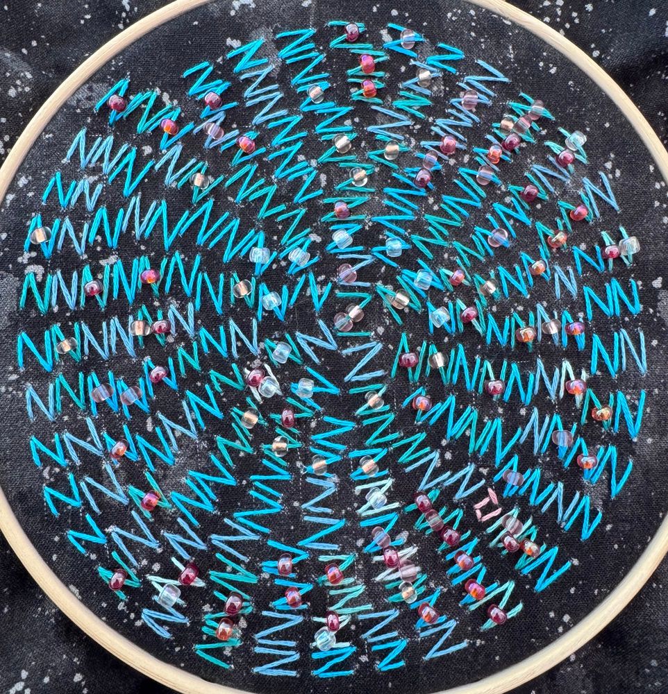 An embroidery hoop of Zs in various shades of blue and teal in a spiral pattern on starry night fabric. A mix of orange, pink, copper, and clear beads are stitched into many of the Zs