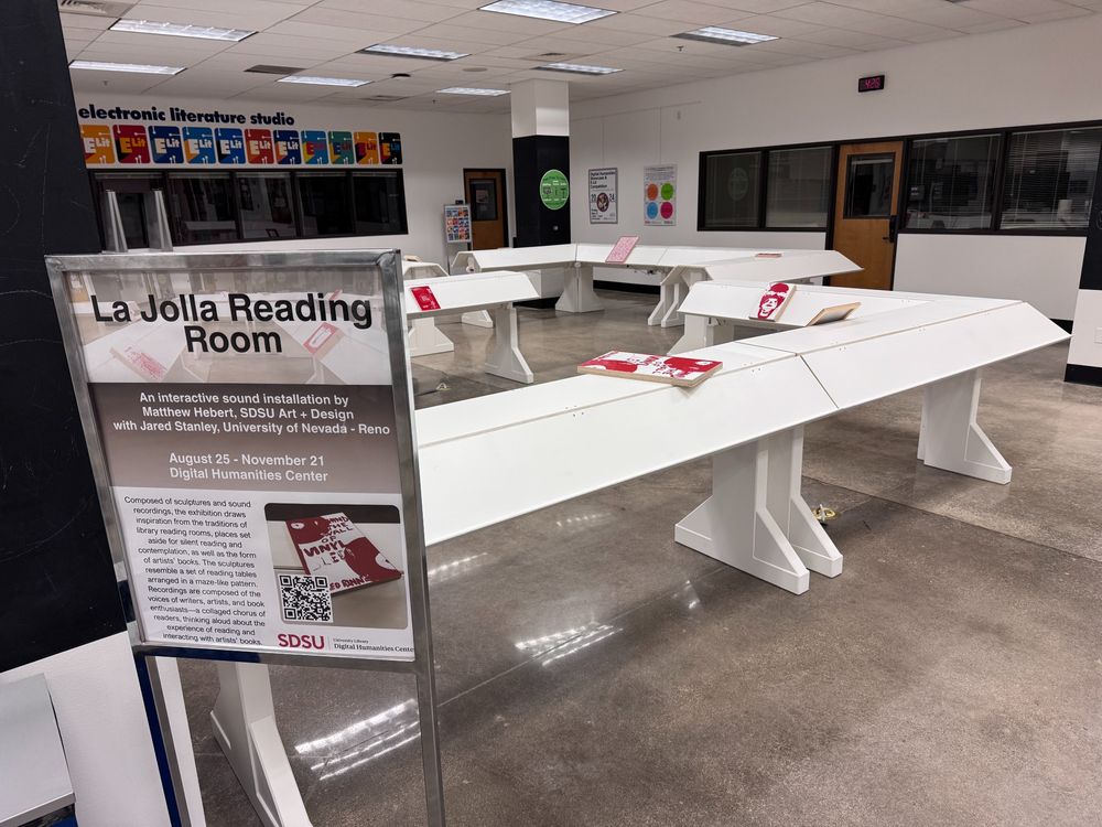 White tables arranged in a maze with red-and-white books fastened to the tables. A sign in a silver stand explains the exhibit just at the start of the installation