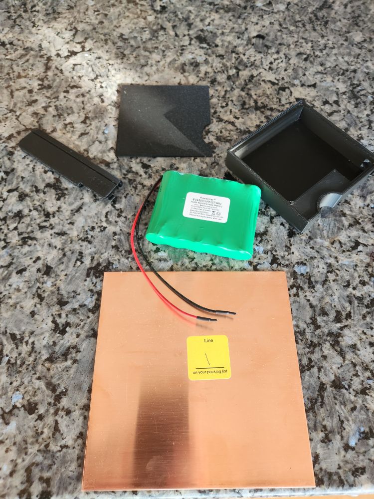 Parts on a granite countertop: 3D printed case components in black, a premade NiMH battery pack with 2 leads, and a sheet of copper.