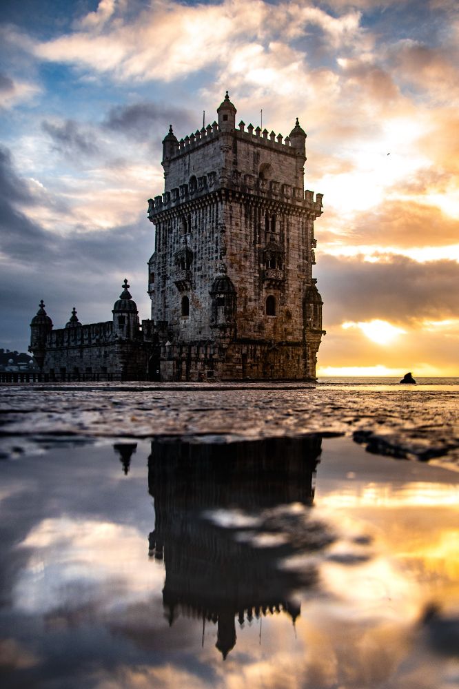 Vue sur la tour de Belém dont on aperçoit le reflet dans une flaque au coucher de Soleil.
Le ciel est chargé est menaçant et les couleurs allant du bleu à l'orange sont très contrastées à cause du contre-jour.