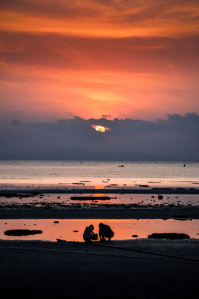 Coucher de Soleil sur une plage thailandaise.
Au premier plan, sur la plage, on aperçoit les silhouettes de deux enfants jouant au bord de l'eau 
Au loin, la mer, et le Soleil couchant sur le point de disparaitre derrière la couche de nuage à l'horizon.
La photo est très contrastée, sombre, et le ciel rougeâtre.