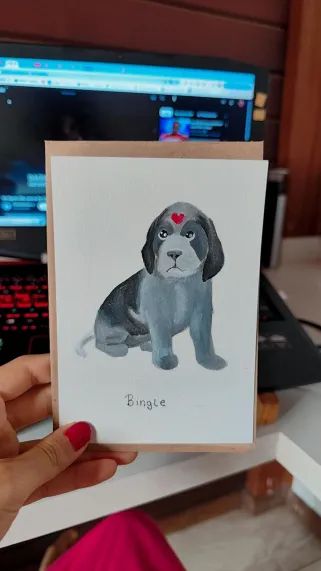 picture of my hand holding a postcard sized painting of a grey and black beagle facing forward with a red heart on its forehead (it's luo binghe)