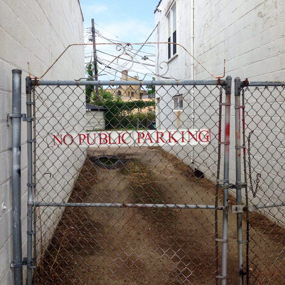 A long white sign with red lettering reads, No public parking. It hangs on a chain link gate in front of an alley between two buildings. 