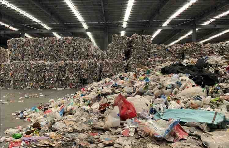 A large warehouse filled with huge stacks of compressed recyclable materials and scattered loose recyclables, creating a sense of industrial scale and environmental urgency.
