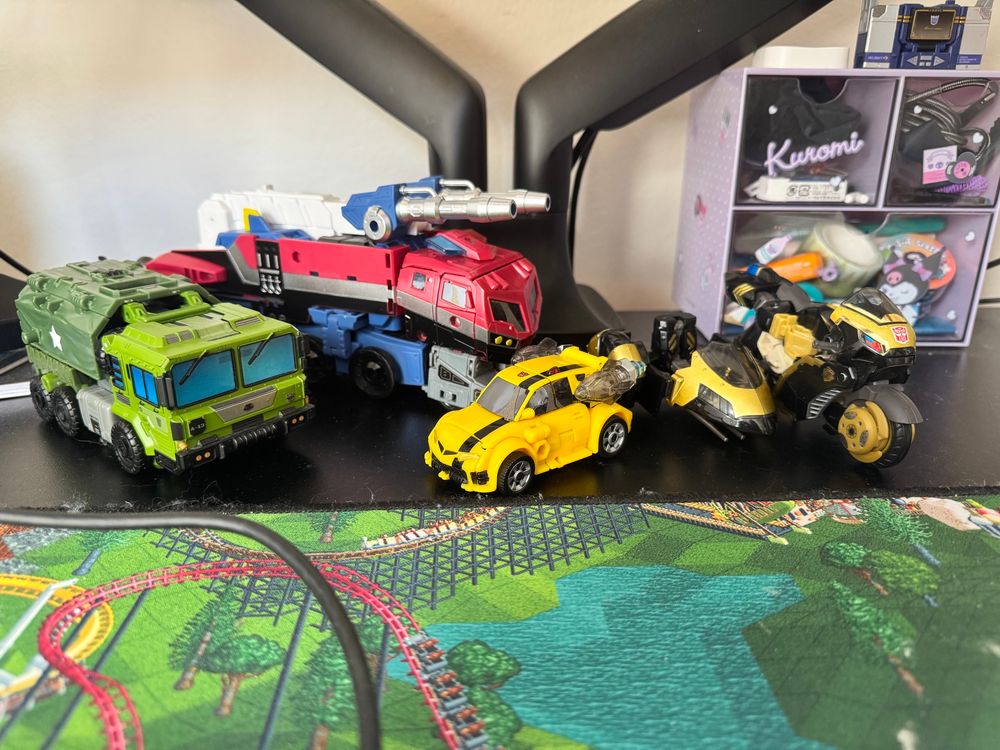 four transformers figures on a desk in vehicle mode. 

from left to right:

legacy voyager prime bulkhead (a large green military transport vehicle with a canopy in the rear)

legacy united voyager animated optimus prime w/ a DNA Designs DK-54 upgrade kit (a red and blue futuristic firetruck with black and silver stripes)

legacy united deluxe animated bumblebee (a small yellow hatchback with a black stripe and rocket boosters)

legacy deluxe animated prowl w/ a DNA Designs DK-42 upgrade kit (a black and gold police motorcycle with a matching sidecar)