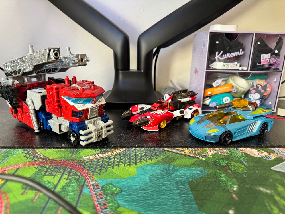 three transformers toys in vehicle mode on a desk. from left to right: seige leader galaxy upgrade optimus prime (a large spacey red and blue firetruck with big cannons), legacy velocitron collection voyager override (a sleek red and white sci-fi race car), and legacy united deluxe cybertron hot shot (a dark tealish countache-inspired sci-fi race car with a winged spoiler and a yellow translucent gun). 